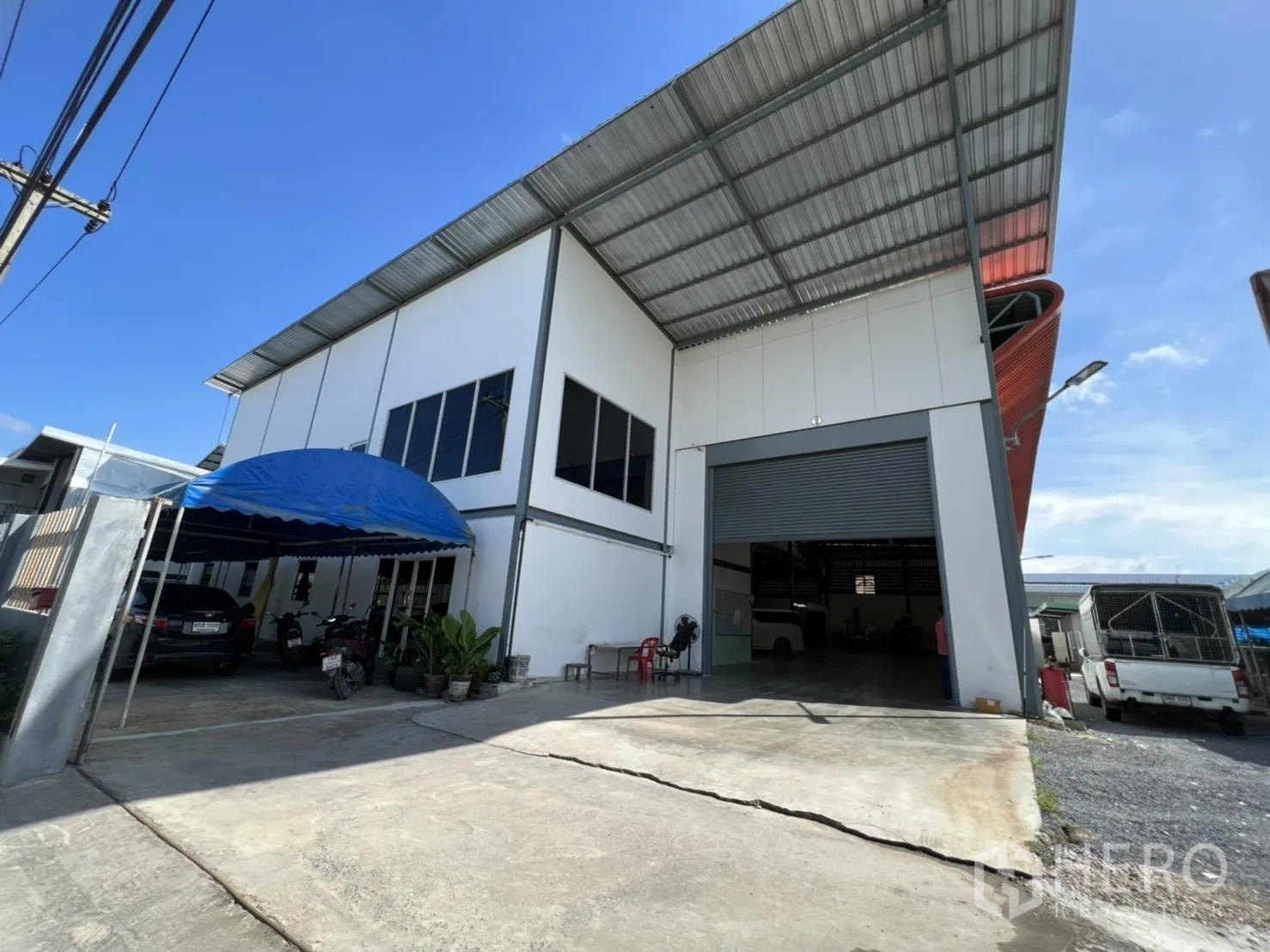 Warehouse for sale in Lam Luk Ka, Pathum Thani - Driveway and wide loading entrance with a blue canopy parking area beside the warehouse facade.