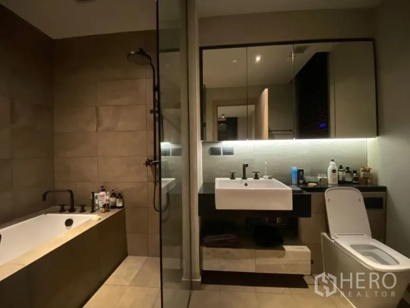 Condominium for rent or sale in Watthana, Bangkok - Luxury bathroom with separate bathtub, glass shower, floating vanity, and warm lighting.