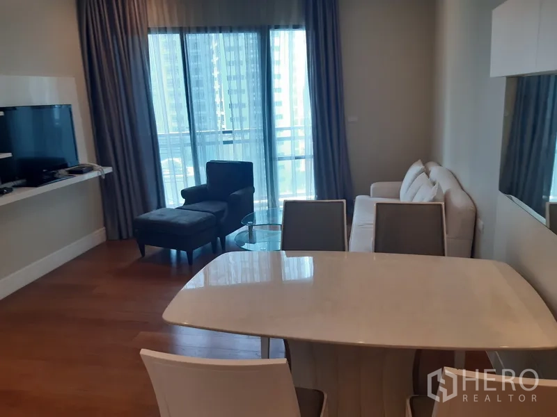 Condominium for rent in Khlong Toei, Bangkok - Bright living and dining area with marble-top table, sofa, TV, and floor-to-ceiling windows to a balcony with city view.
