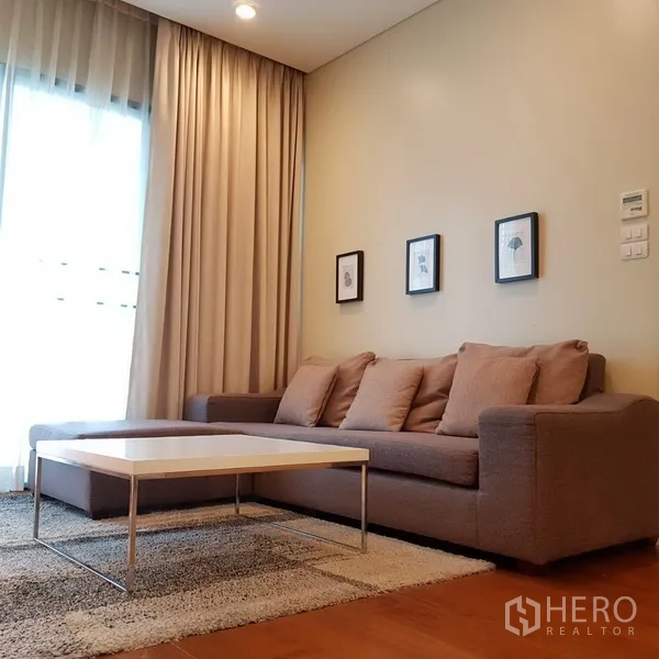 Condominium for rent in Khlong Toei, Bangkok - Living area with gray sectional sofa, coffee table and large curtains bringing in soft light.