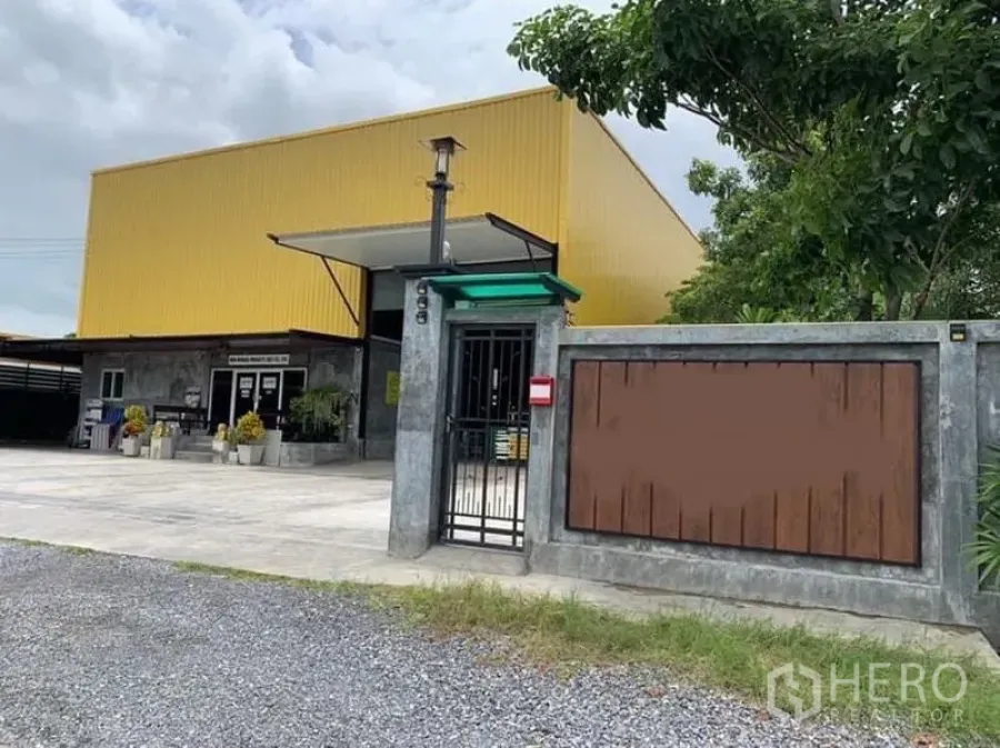 工厂 出售在Thanyaburi, Pathum Thani - 带大门与围墙的黄色外墙工厂入口，前方为宽阔混凝土场地与绿树。