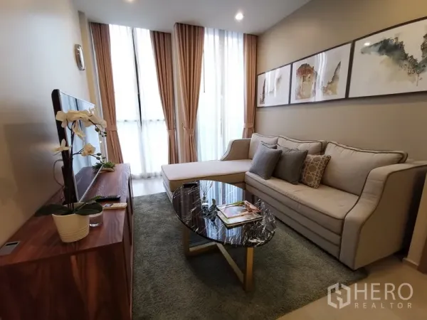Condominium for rent in Pathum Wan, Bangkok - Cozy living room with L-shaped sofa, wall art, and tall curtained windows.