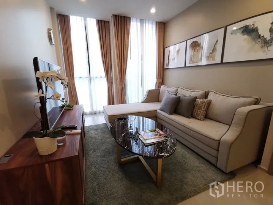 คอนโด ให้เช่าในPathum Wan, Bangkok - ห้องนั่งเล่นอบอุ่น โซฟา L‑shape งานศิลป์บนผนัง และหน้าต่างสูงพร้อมผ้าม่าน