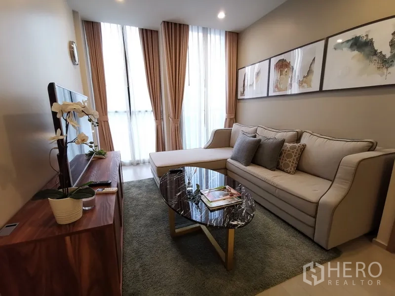 คอนโด ให้เช่าในPathum Wan, Bangkok - ห้องนั่งเล่นอบอุ่น โซฟา L‑shape งานศิลป์บนผนัง และหน้าต่างสูงพร้อมผ้าม่าน