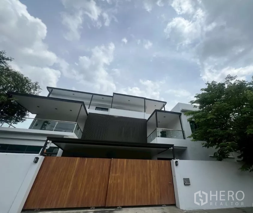 带办公室的家 出租在Watthana, Bangkok - 现代多层家用办公楼外观，白色立面与玻璃阳台，宽木质大门，阴云天空下