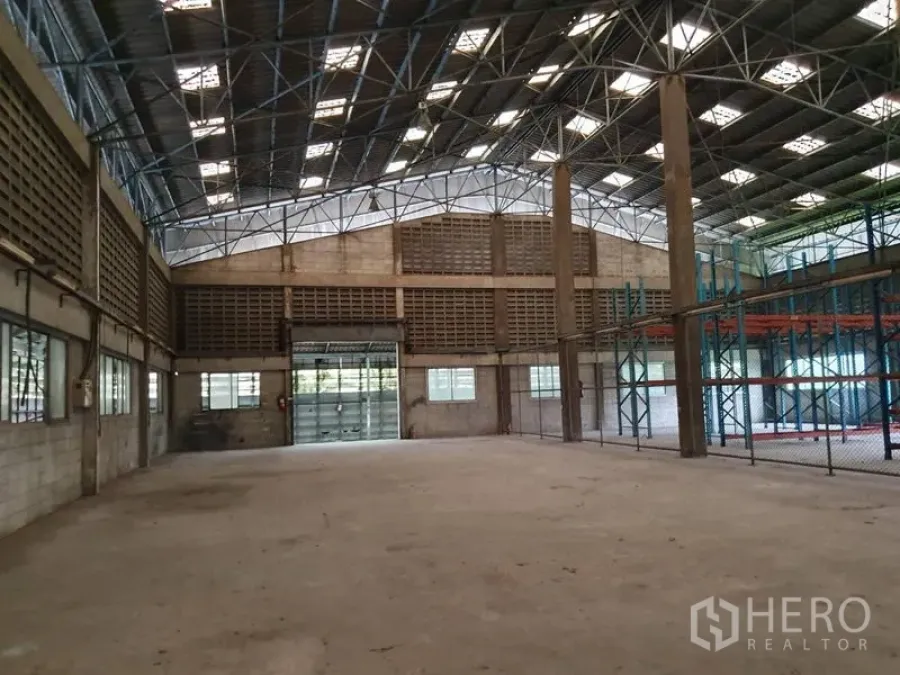 Factory for rent in Samut Prakan - Spacious factory hall with a high steel-truss roof, skylights, ventilation block walls and a large roller shutter door.