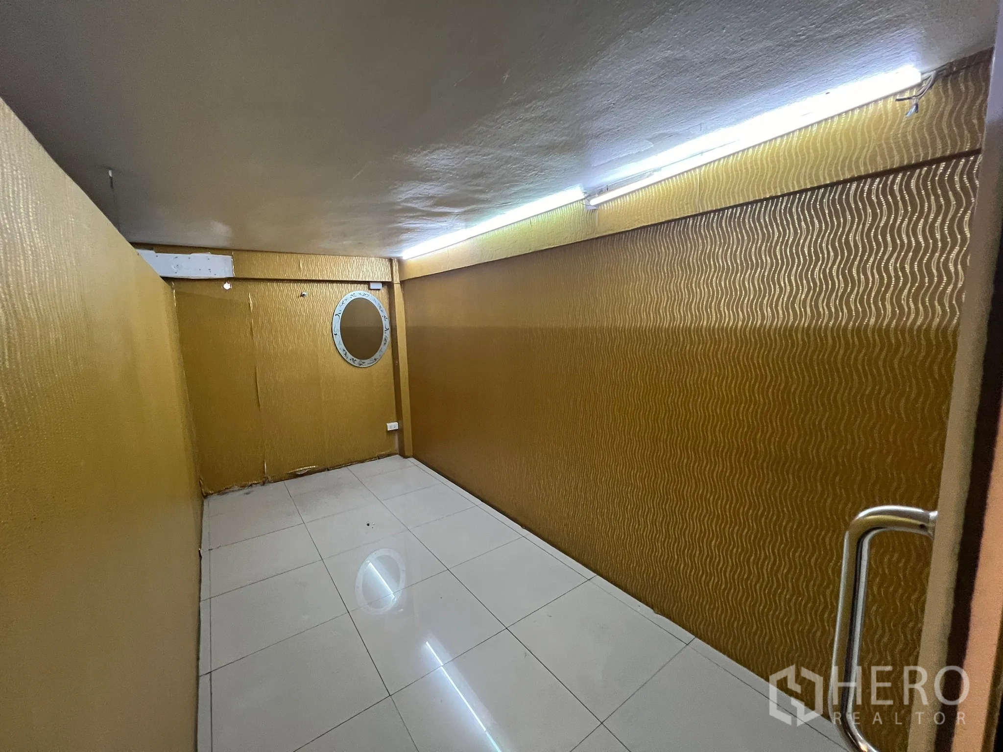 Shophouse for rent in Khlong Toei, Bangkok - Long gold-toned room with fluorescent lighting and a round mirror on the wall.