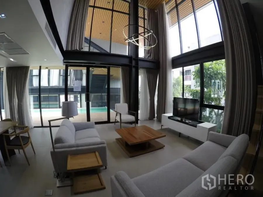 Apartment for rent in Khlong Toei, Bangkok - Double-height living room with large glazing, grey sofas, and outlook to the pool terrace.