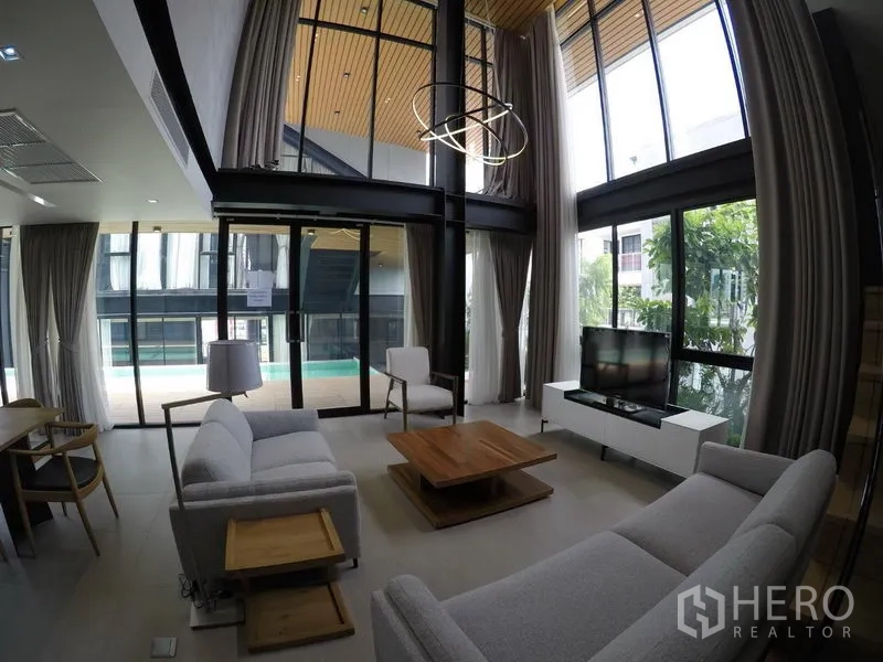 Apartment for rent in Khlong Toei, Bangkok - Double-height living room with large glazing, grey sofas, and outlook to the pool terrace.