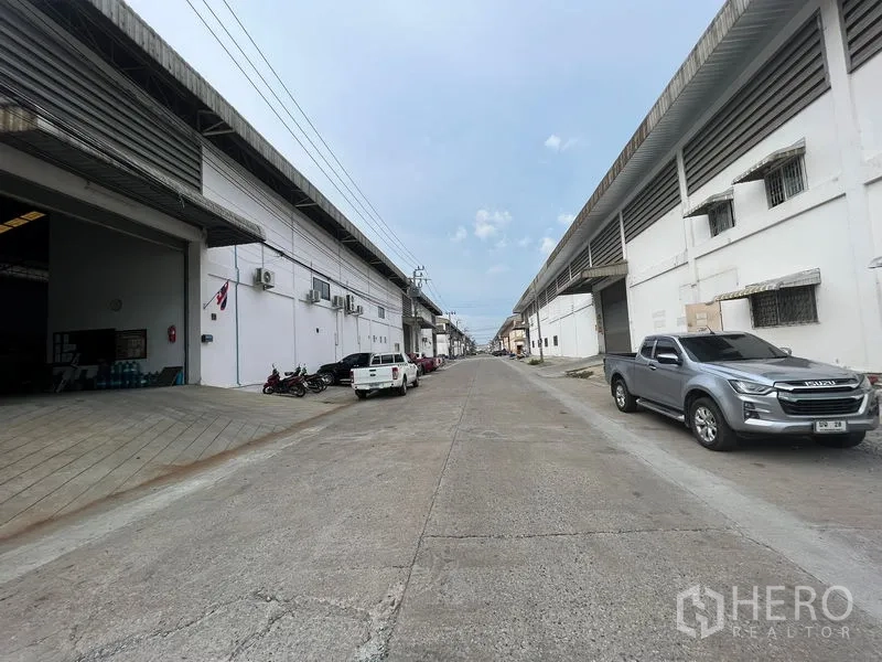 货栈 出租在Bang Sao Thong, Samut Prakan - 工业区车道，两侧为仓库建筑，停有皮卡车。