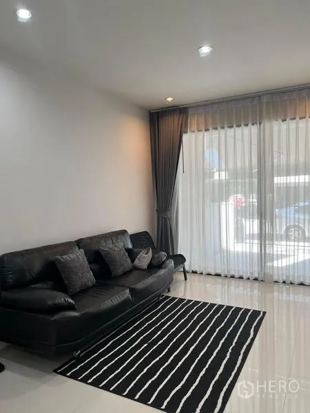 Townhouse for rent in Saphan Sung, Bangkok - Living room with black leather sofa, striped rug and floor‑to‑ceiling sheer curtains.