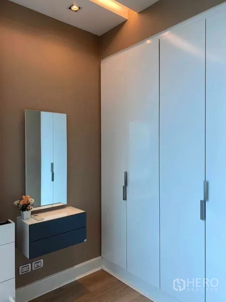 Condominium for rent in Bangkok - Bedroom area with large built-in white wardrobe and wall mirror over a floating cabinet.