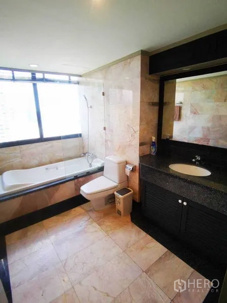 Condominium for rent in Khlong Toei, Bangkok - Marble-tiled bathroom with bathtub behind glass screen, toilet and wide vanity mirror.