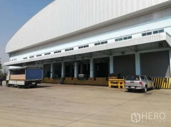 Warehouse for rent in Nong Chok, Bangkok - Row of covered loading docks with roll-up doors and trucks parked on a wide concrete apron.