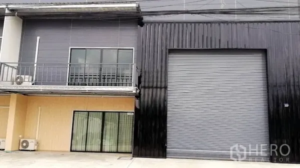Warehouse for rent or sale in Lam Luk Ka, Pathum Thani - Close view of the warehouse facade showing a tall roller door, glass-fronted office with balcony, and exterior AC units.