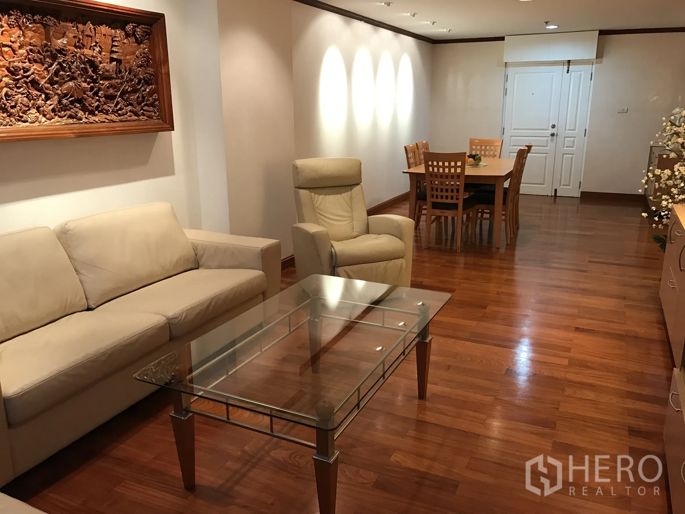 Condominium for rent in Khlong Toei, Bangkok - Living area facing dining table with wood floors and carved wall art.