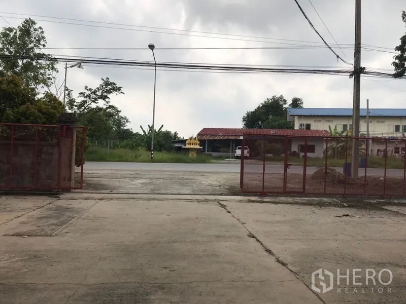 Warehouse for rent in Mueang Nakhon Pathom, Nakhon Pathom - Gated warehouse entrance opening to a main road with a streetlight and neighboring buildings.