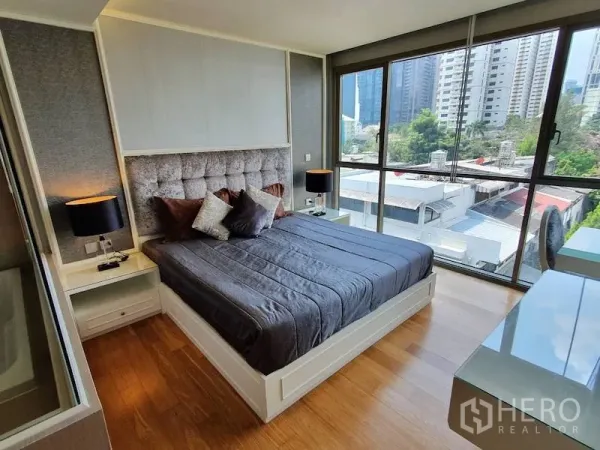 公寓 出租在Watthana, Bangkok - 转角卧室配落地窗、软包床头与床头灯，木地板与城市景观。