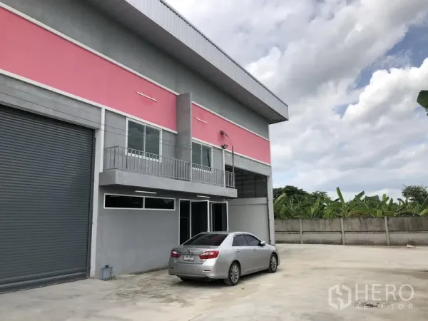 Factory for rent in Sai Noi, Nonthaburi - Corner unit showing a roller shutter door, balcony and generous parking apron beside the boundary wall.