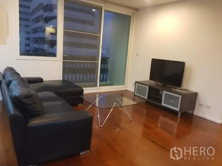 公寓 出租在Watthana, Bangkok - 客厅配黑色皮沙发、玻璃茶几和电视柜，木地板，推拉门通往可观城市景观的阳台。