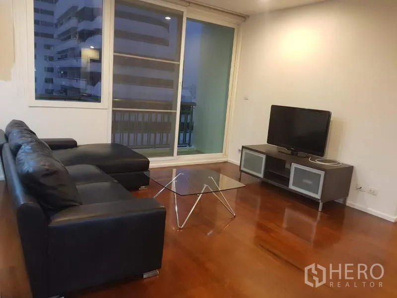 公寓 出租在Watthana, Bangkok - 客厅配黑色皮沙发、玻璃茶几和电视柜，木地板，推拉门通往可观城市景观的阳台。