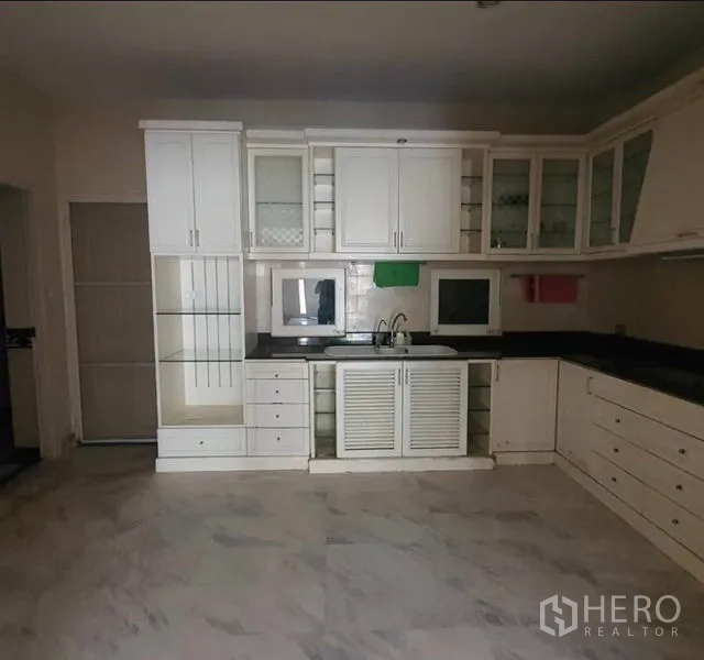 Townhouse for rent in Huai Khwang, Bangkok - White L-shaped kitchen with glass-front cabinets, double sink and black countertops.