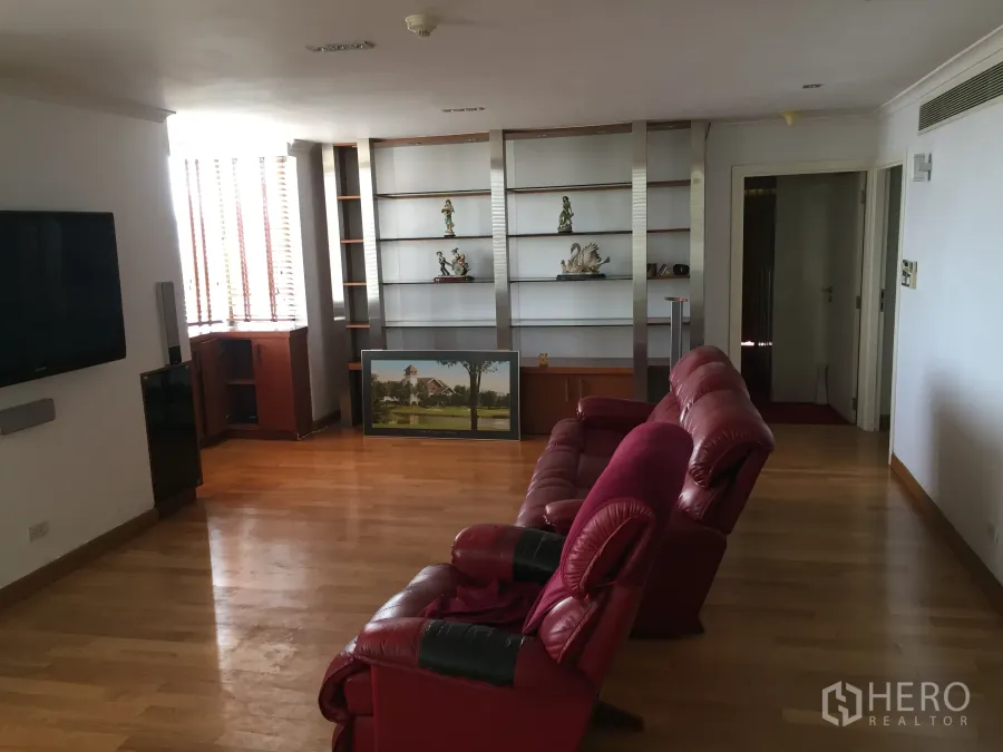 Condominium for rent in Khlong Toei, Bangkok - Spacious living room with wood floors, red leather recliners, built-in shelving and a wall-mounted TV.