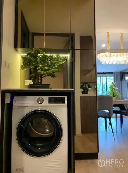 公寓 出租在Pathum Wan, Bangkok - 镜面柜下嵌入式滚筒洗衣机，可见餐区与吊灯景致。