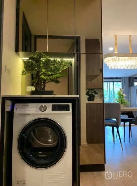 公寓 出租在Pathum Wan, Bangkok - 镜面柜下嵌入式滚筒洗衣机，可见餐区与吊灯景致。