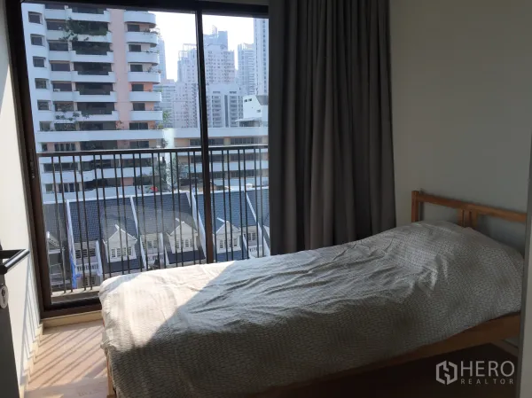 Condominium for rent in Bangkok - Sunlit bedroom with single bed and sliding glass doors to a balcony overlooking city buildings.