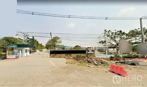 Factory for sale in Lam Luk Ka, Pathum Thani - Gated industrial entrance in Lam Luk Ka with security booth, boundary wall and wide access road under power lines.