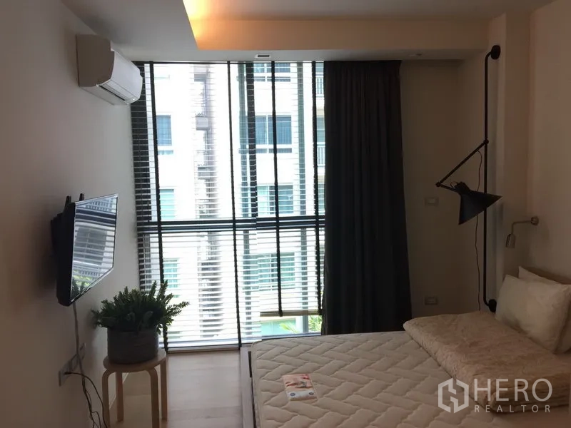 Condominium for rent in Watthana, Bangkok - Bedroom with floor-to-ceiling windows, blinds, wall TV and indoor plant beside the bed.