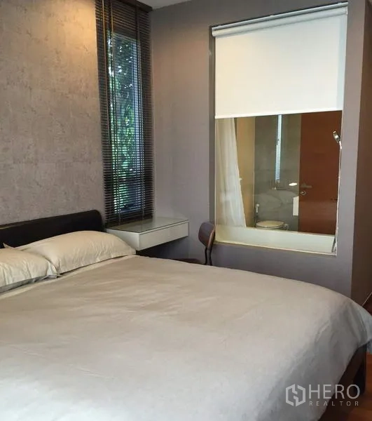 Condominium for rent in Khlong Toei, Bangkok - Cozy bedroom with king bed, window to ensuite bathroom with roller blind, and side window with blinds facing greenery.