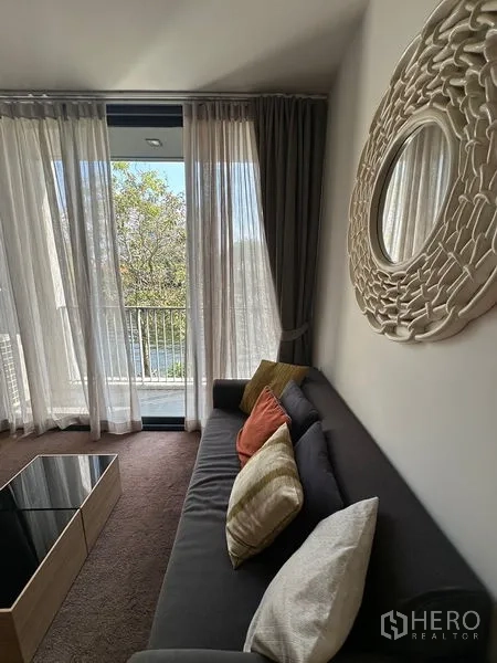 Condominium for rent in Watthana, Bangkok - Living room sofa beside balcony doors with sheer curtains and decorative round mirror.
