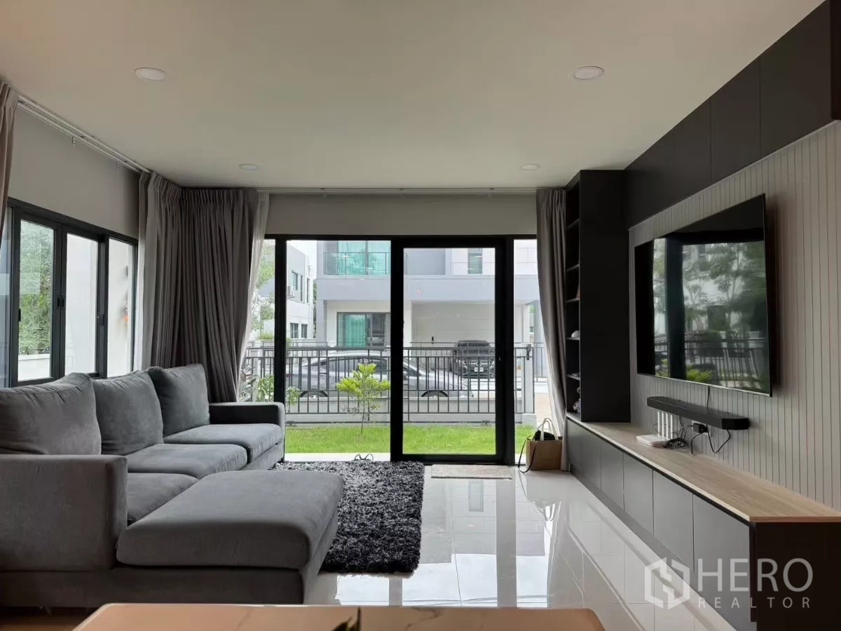 独立式住宅 出租在Bang Phli, Samut Prakan - 明亮客厅，灰色转角沙发、电视墙，推拉门通向花园。