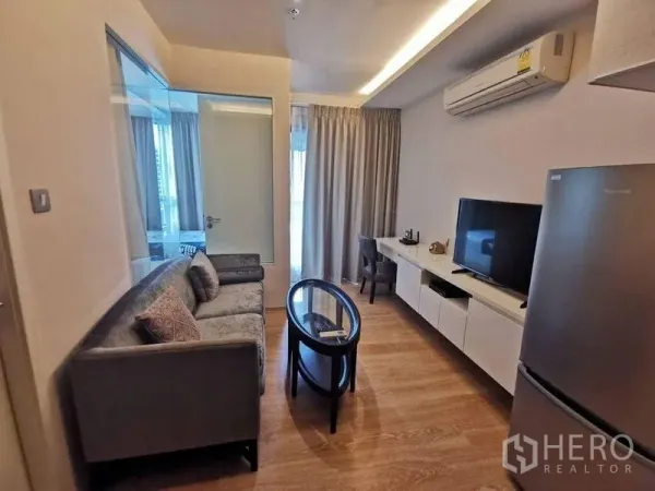 公寓 出租或出售在Watthana, Bangkok - 客厅配灰色沙发、玻璃茶几、电视与书桌，靠近大窗帘