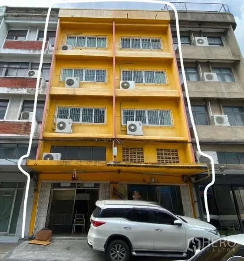 Shophouse for rent or sale in Watthana, Bangkok - Yellow multi-storey shophouse facade with multiple A/C units and ground-level parking.