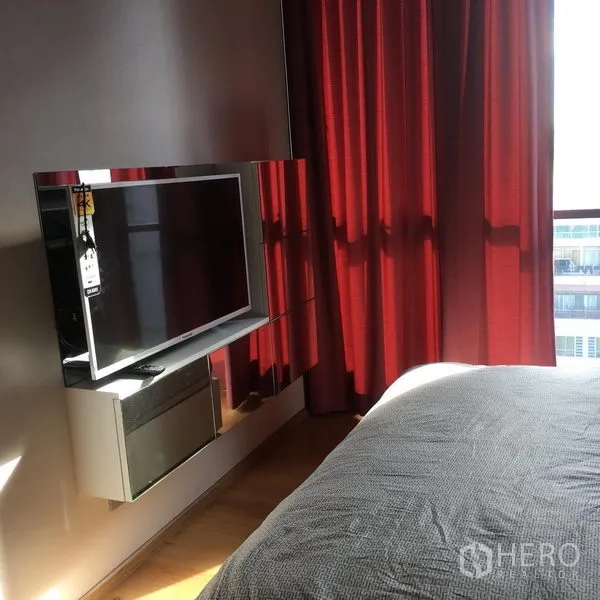 Condominium for rent or sale in Watthana, Bangkok - Bedroom corner with wall-mounted TV, red blackout curtains and natural light.