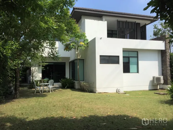 独立式住宅 出租或出售在Prawet, Bangkok - 后花园草坪与成树，白色立面旁的庭院桌椅和推拉落地门。