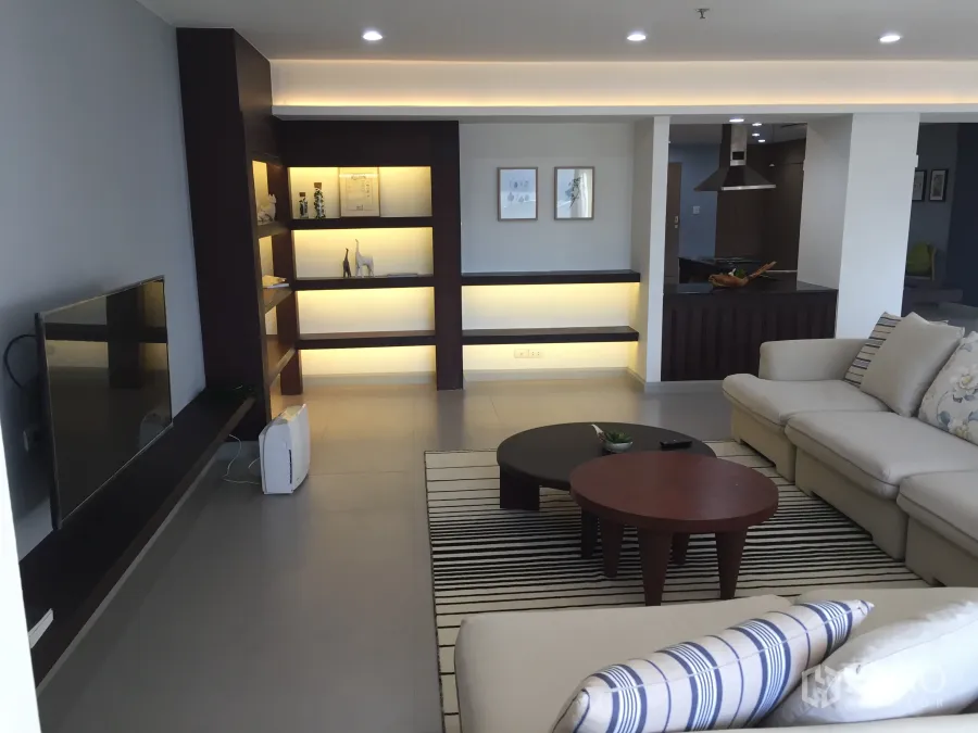 Condominium for sale in Watthana, Bangkok - Spacious living room with backlit built-in shelves, sectional sofa, round coffee tables, TV wall and recessed lighting beside an open kitchen.