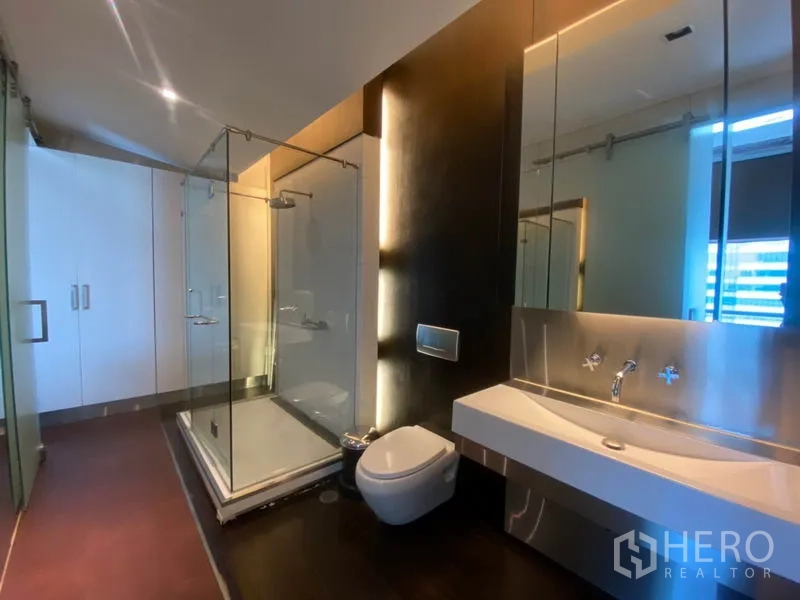 Condominium for rent in Sathon, Bangkok - Modern bathroom featuring a glass-enclosed shower, wall-hung toilet and long vanity with mirror lighting.