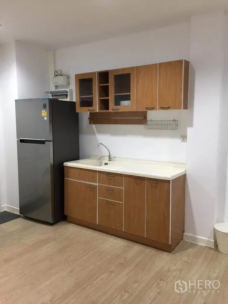 Townhouse for rent in Lat Phrao, Bangkok - Kitchen corner with upper and lower cabinets, sink and stainless refrigerator on wood floors.