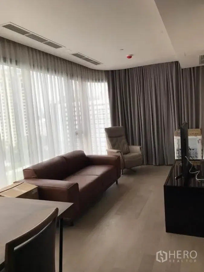 Condominium for rent in Watthana, Bangkok - Bright living and dining area with floor‑to‑ceiling windows, sheer curtains, brown leather sofa and TV console overlooking the city.