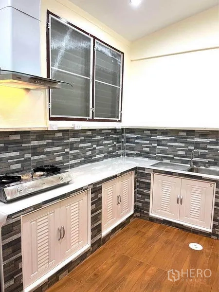 Townhouse for rent in Bang Na, Bangkok - L-shaped kitchen with sink, white cabinets, gray mosaic backsplash and wood-look floor.