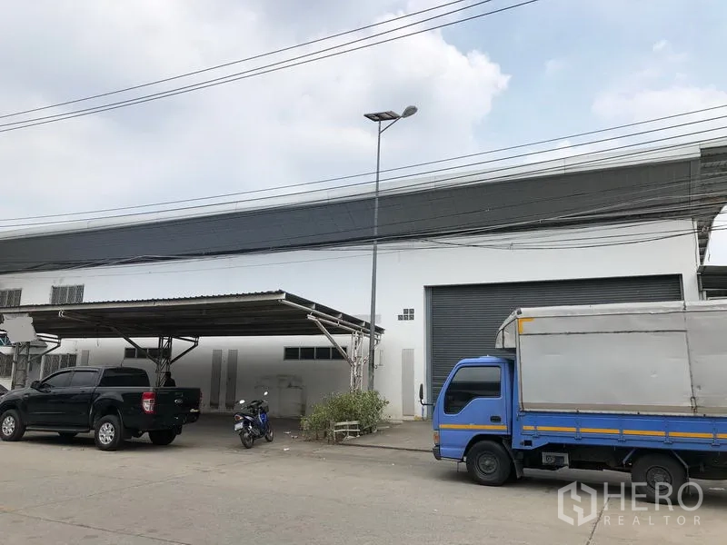 Factory for rent in Nonthaburi - Angled exterior of the warehouse showing a covered carport and gray roller door with a streetlight.