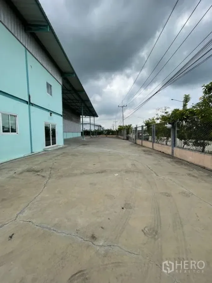 货栈 出租在Lat Lum Kaeo, Pathum Thani - 仓库外部车道，带雨棚装卸区、围栏与电线，天空多云。