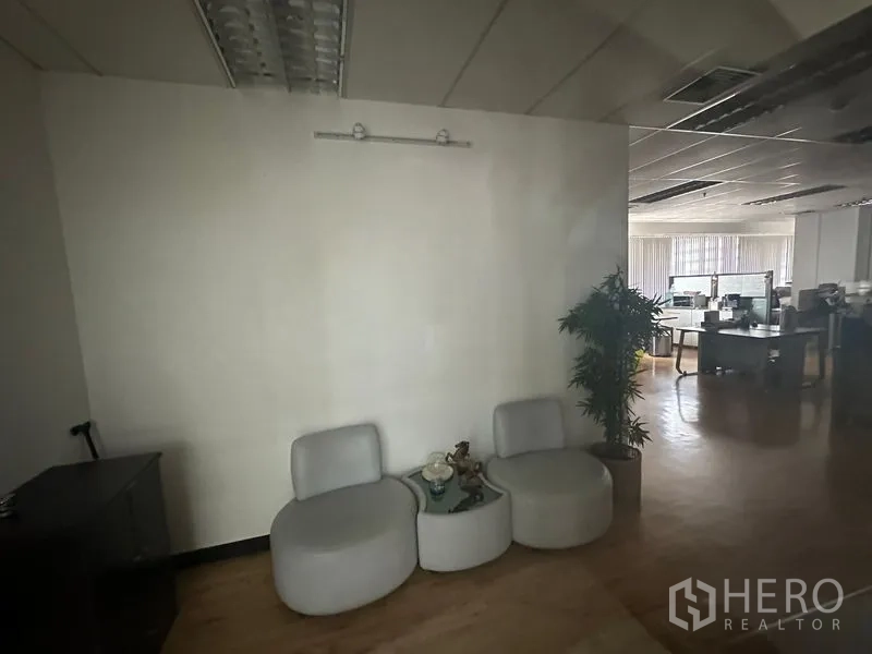 Office Space for rent in Bang Rak, Bangkok - Reception nook with two gray lounge chairs, a small round table, an indoor plant, and a view to an open-plan office with desks and blinds.