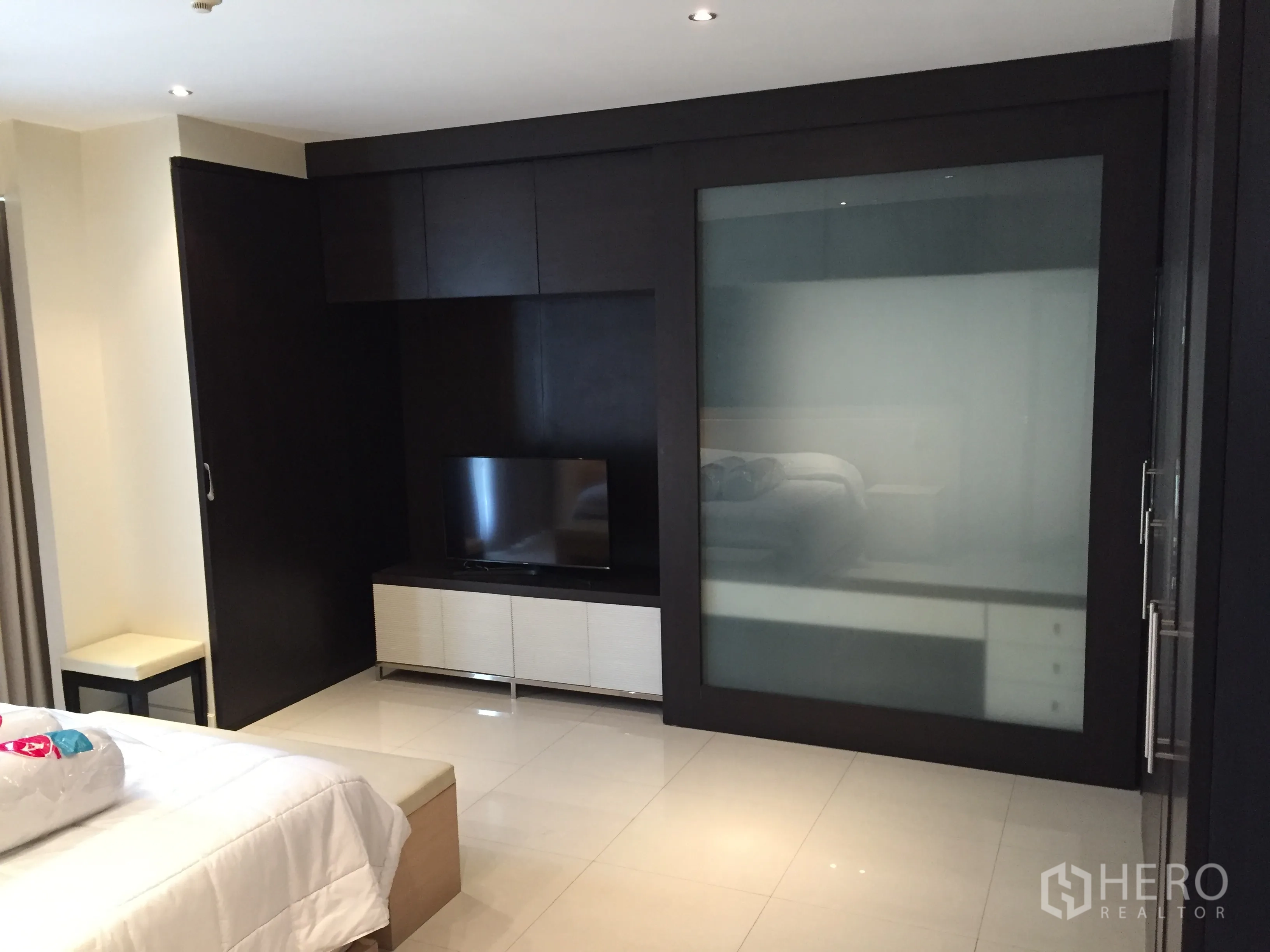 Condominium for rent in Bang Rak, Bangkok - Bright bedroom with built-in TV wall, frosted-glass wardrobe and glossy tile floors.