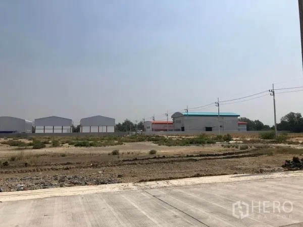 Land for sale in Mueang Samut Sakhon, Samut Sakhon - Vacant industrial land with a concrete road, power lines, and nearby warehouse buildings in Samut Sakhon.