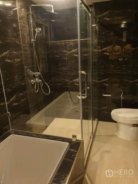 Condominium for rent in Khlong Toei, Bangkok - Modern bathroom with glass shower, dark marble tiles, bathtub and toilet.